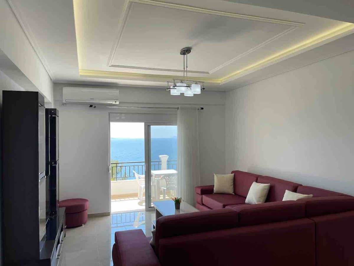 A modern living area features a spacious red sectional sofa arranged around a sleek coffee table. Large glass doors provide access to a balcony with ocean views, while a contemporary light fixture hangs from the ceiling. A soft neutral palette enhances the airy ambiance.