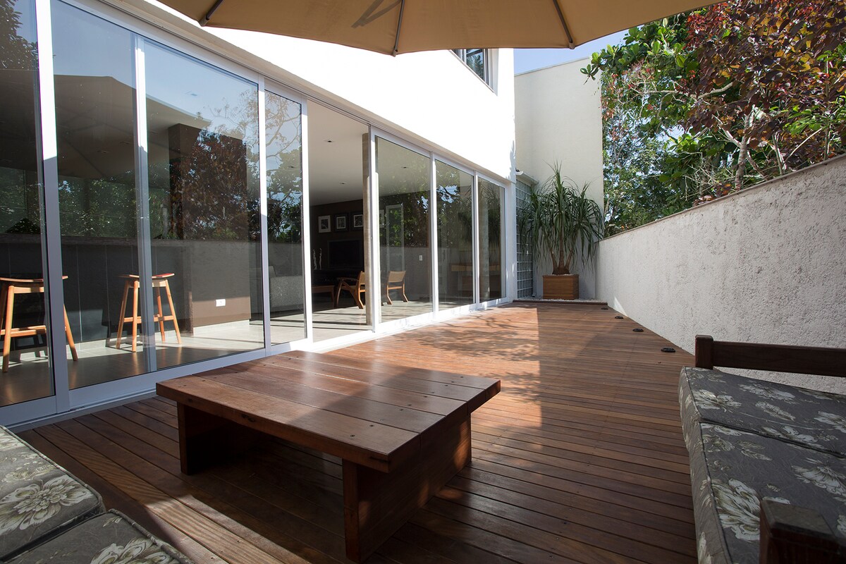 A spacious outdoor deck is featured, with wooden flooring and a low table at the center. Large glass sliding doors offer a view into the interior. The area is shaded by a canopy, surrounded by greenery and modern furnishings, creating an inviting environment.