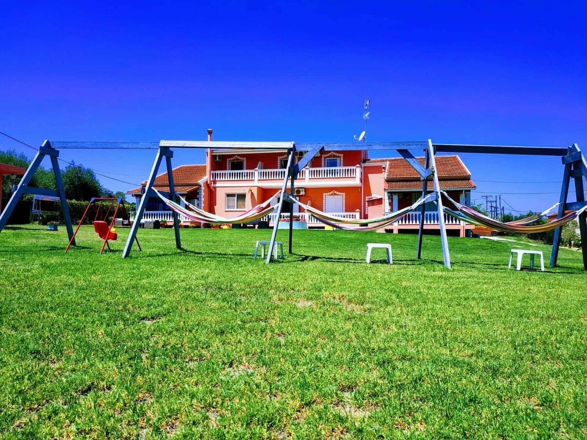 A spacious outdoor area is featured, showcasing several hammocks suspended between sturdy frames on a well-maintained lawn. In the background, a charming multi-story building with a pink facade and balconies is visible, under a clear blue sky.