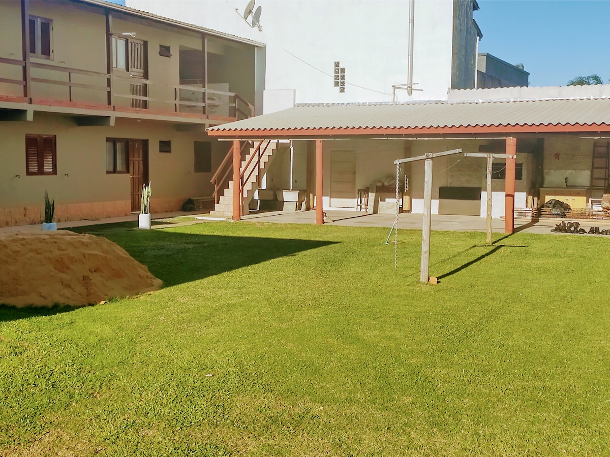 A spacious shared outdoor area features well-maintained green grass and a relaxing atmosphere. A covered patio area is visible in the background, while a playground swing set stands on one side. The layout is open, inviting leisure and family gatherings.