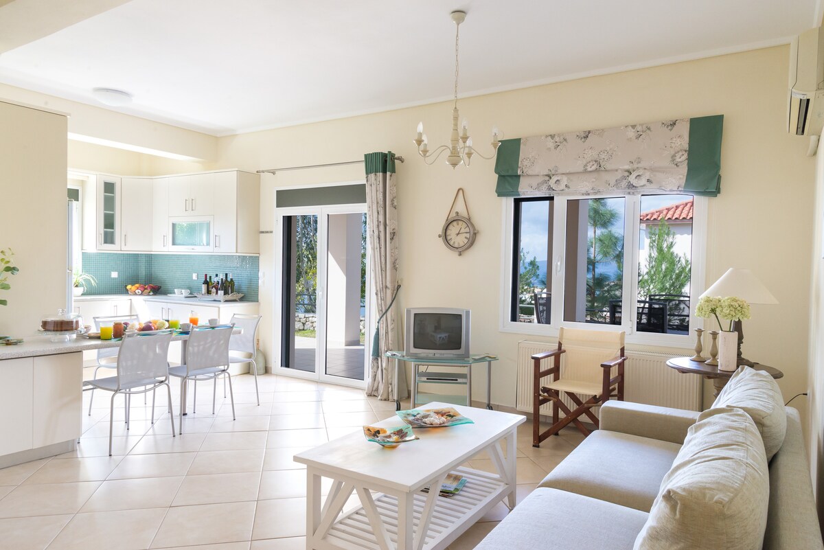 A spacious living area is shown, with light-colored tiled floors and large windows allowing natural light. A dining table with chairs is positioned next to the kitchen, which features modern cabinets and appliances. A comfortable sofa sits beside a coffee table, complemented by decor items.