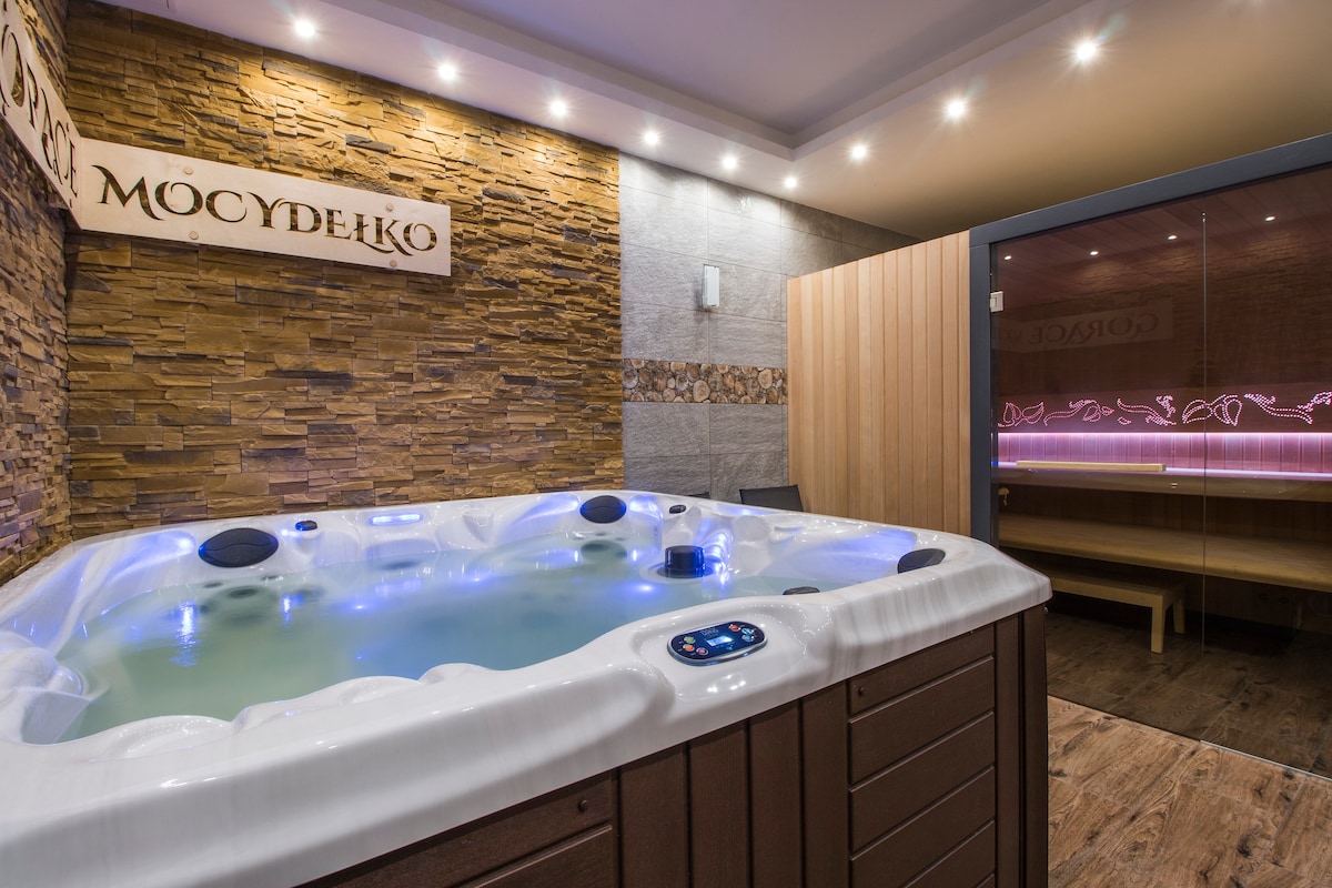A well-lit area features a spacious jacuzzi with soft blue lighting, surrounded by a stone accent wall. Adjacent, a sauna with wooden paneling and glass doors is visible, inviting relaxation and wellness.