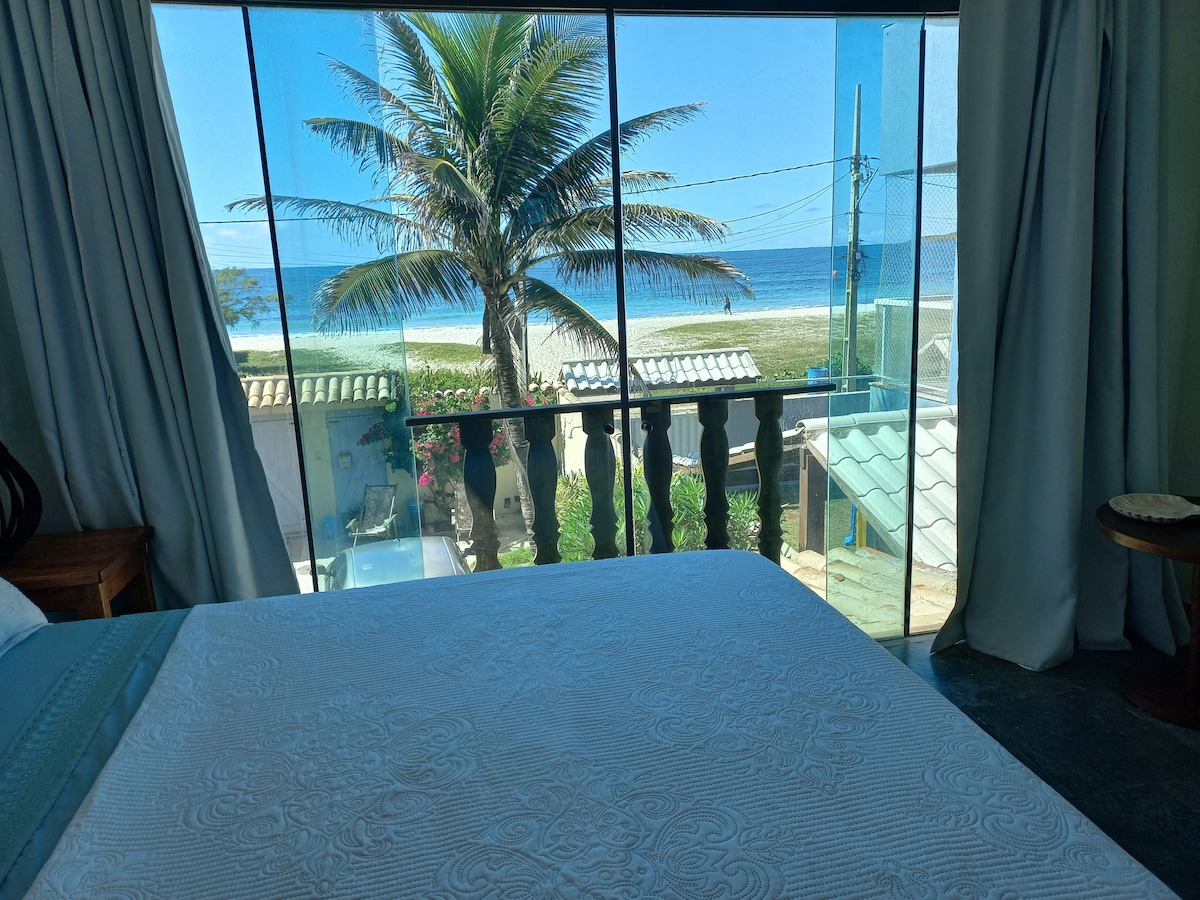 A bright bedroom features a spacious bed positioned for optimal ocean views. Large windows reveal a clear view of the beach and palm trees outside. Soft curtains frame the window, allowing natural light to fill the space.