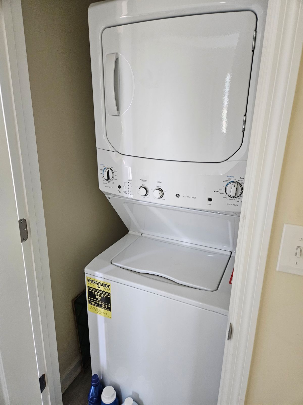 A stacked washer and dryer unit is positioned in a narrow space. The white exterior features dials and controls on the top portion, with laundry detergent visible at the base. A door partially opens to the area, maximizing efficiency in laundry tasks.