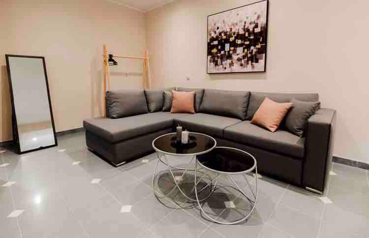A spacious living area showcases a large, grey sectional sofa adorned with decorative pillows. A dark glass coffee table is positioned in front, accompanied by two minimalist circular tables. A full-length mirror reflects the light from the room, enhancing the overall ambiance.