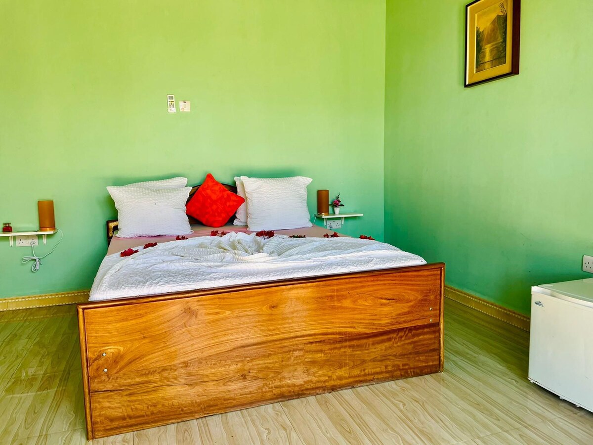 A spacious bed is centered in a room with vibrant green walls, featuring decorative red accents. Nightstands are positioned on either side, and a small fridge is located to the right. Soft light fills the space, highlighting the warm wooden textures.