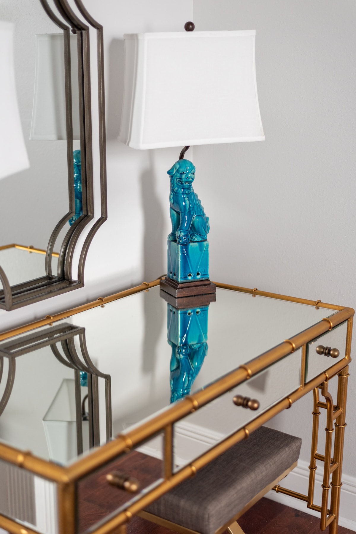 A stylish brass-framed vanity table is featured, topped with a mirrored surface reflecting a bright blue lamp. The lamp's base is shaped like a figure, adding a whimsical touch. A neutral upholstered chair sits nearby, complementing the elegant design.