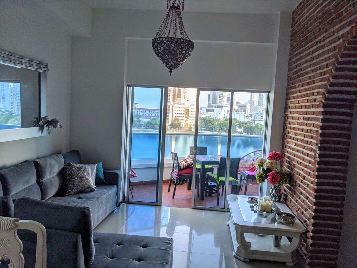 A modern living area is visible, featuring a comfortable gray sofa adorned with decorative pillows. Large glass doors open to a scenic view of the water, and a dining table is set with colorful chairs and a floral centerpiece.