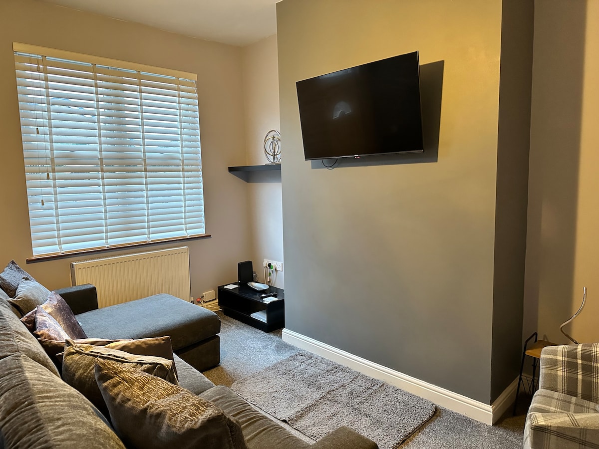 A comfortable living area is presented, featuring a large sectional sofa with decorative pillows. A flat-screen television is mounted on the wall, above a shelf supporting media devices. Light filtered through the window highlights the neutral tones and soft textures of the carpet.