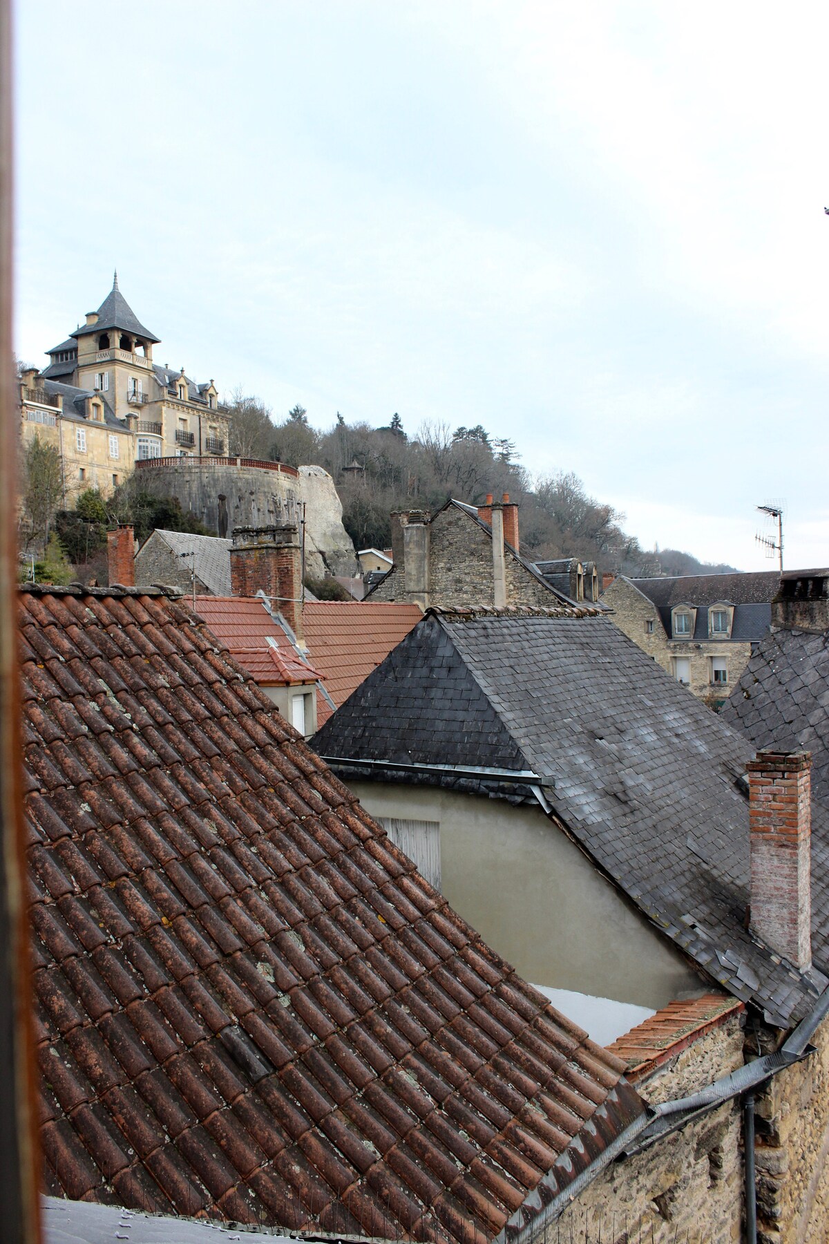 A scenic view of the historic rooftops of Montignac is presented, showcasing a mix of slate and terracotta tiles. Above the rooftops, a castle with a distinctive turret is visible, set against a backdrop of trees and a cloudy sky.