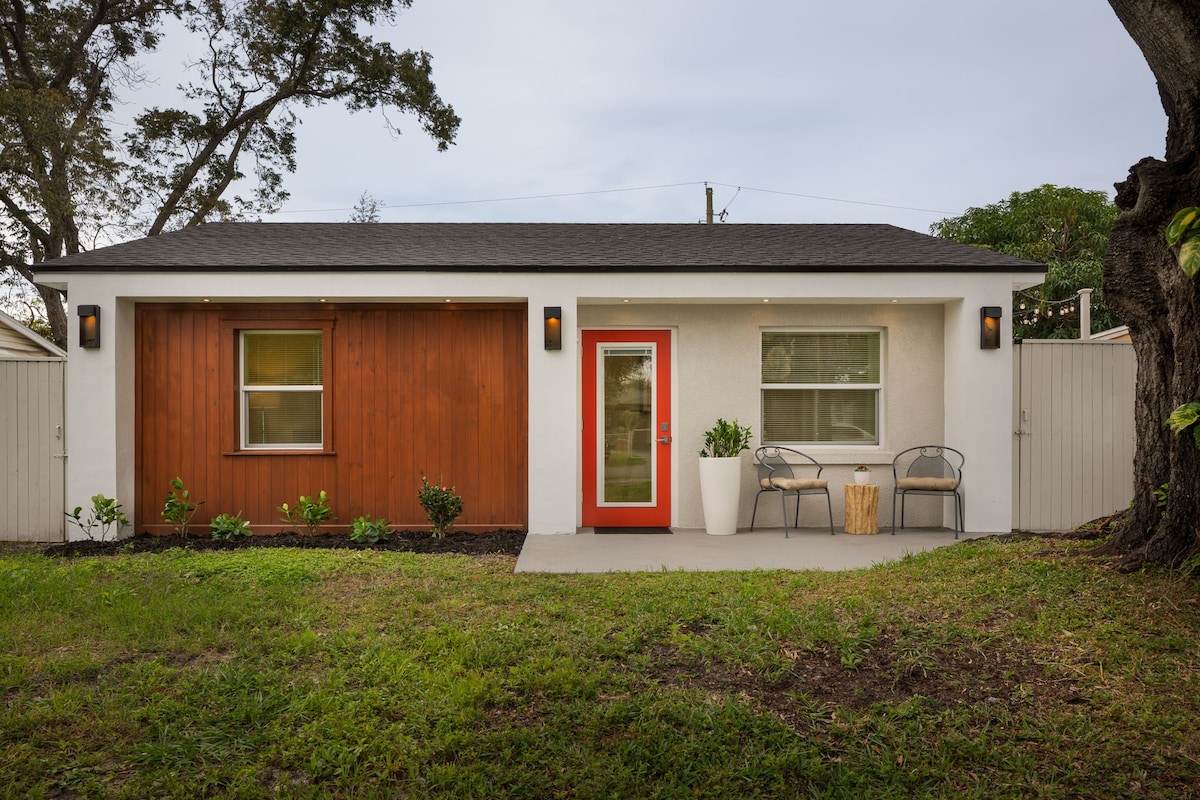 A modern cottage exterior features two contrasting wall materials, one side clad in wood and the other in smooth white stucco. A bright red door serves as an inviting focal point. A small seating area with gray chairs is positioned on the porch, surrounded by lush greenery.