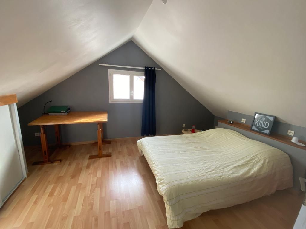 A bright bedroom is featured under a pitched ceiling, with a spacious bed and a wooden table positioned by the window. Soft lighting highlights the light-colored flooring, while a dark curtain complements the white walls.
