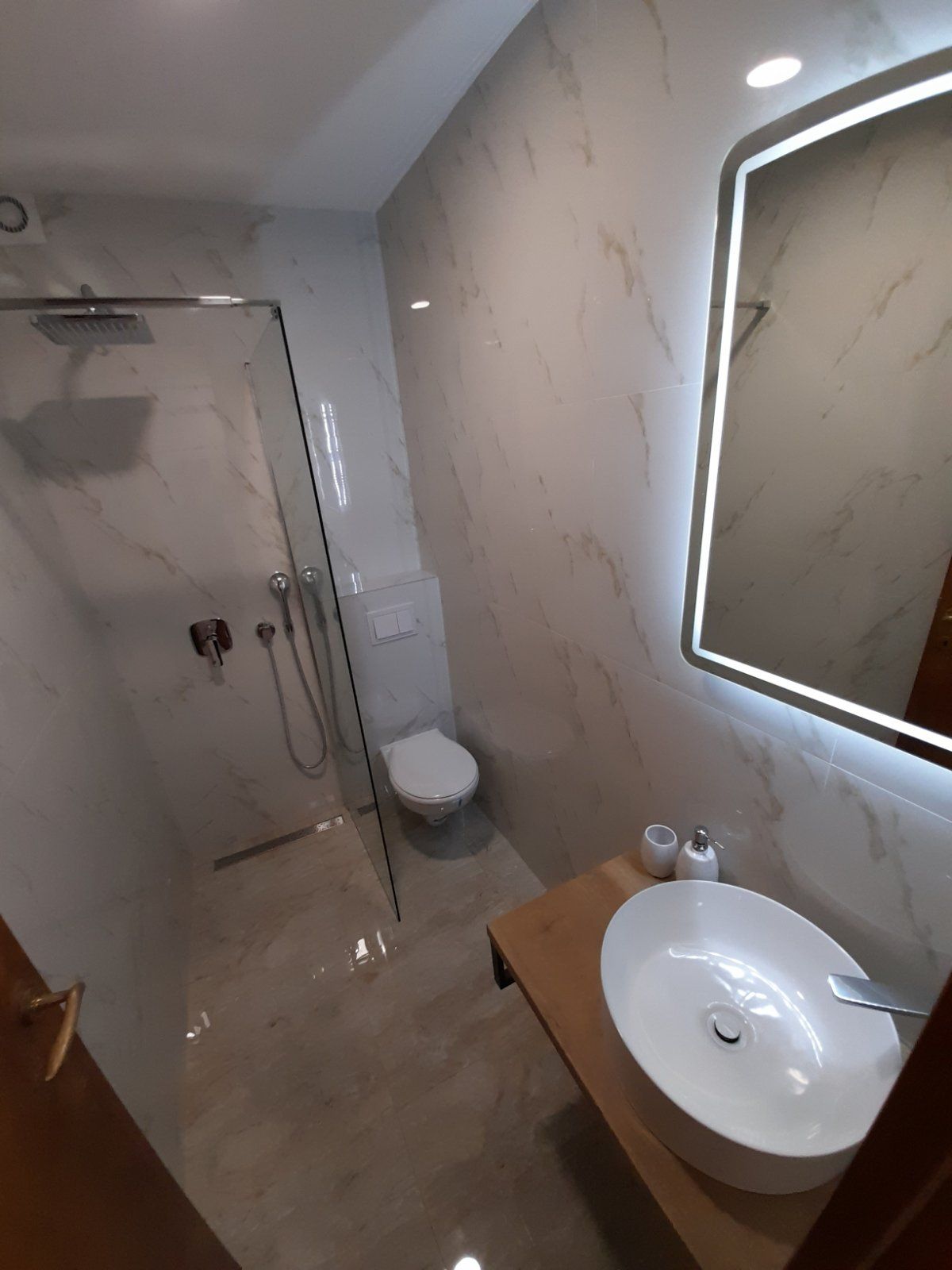 A modern bathroom is presented with sleek marble walls and a glass shower enclosure. A circular mirror with integrated lighting hangs above a contemporary sink on a wooden countertop. A toilet is positioned discretely, complementing the elegant and spacious layout.