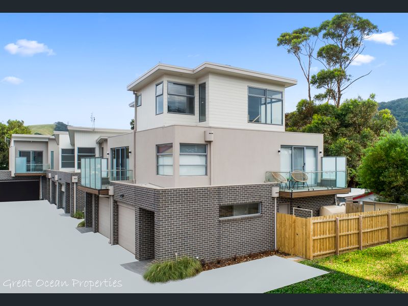 A modern townhouse exterior is showcased with clean lines and a mix of materials, featuring large glass balcony railings. Lush greenery surrounds the property, and a wooden fence provides privacy. Driveways are visible leading to garages, enhancing the functional design of the home.