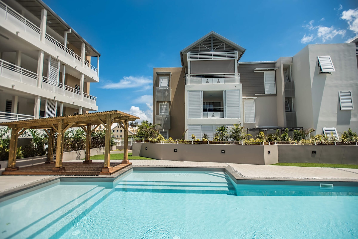 A modern residence is shown, featuring light-colored exteriors and a spacious swimming pool in the foreground. Lush landscaping surrounds the pool area, and a wooden gazebo is present nearby. Bright blue skies enhance the relaxation setting of the outdoor space.