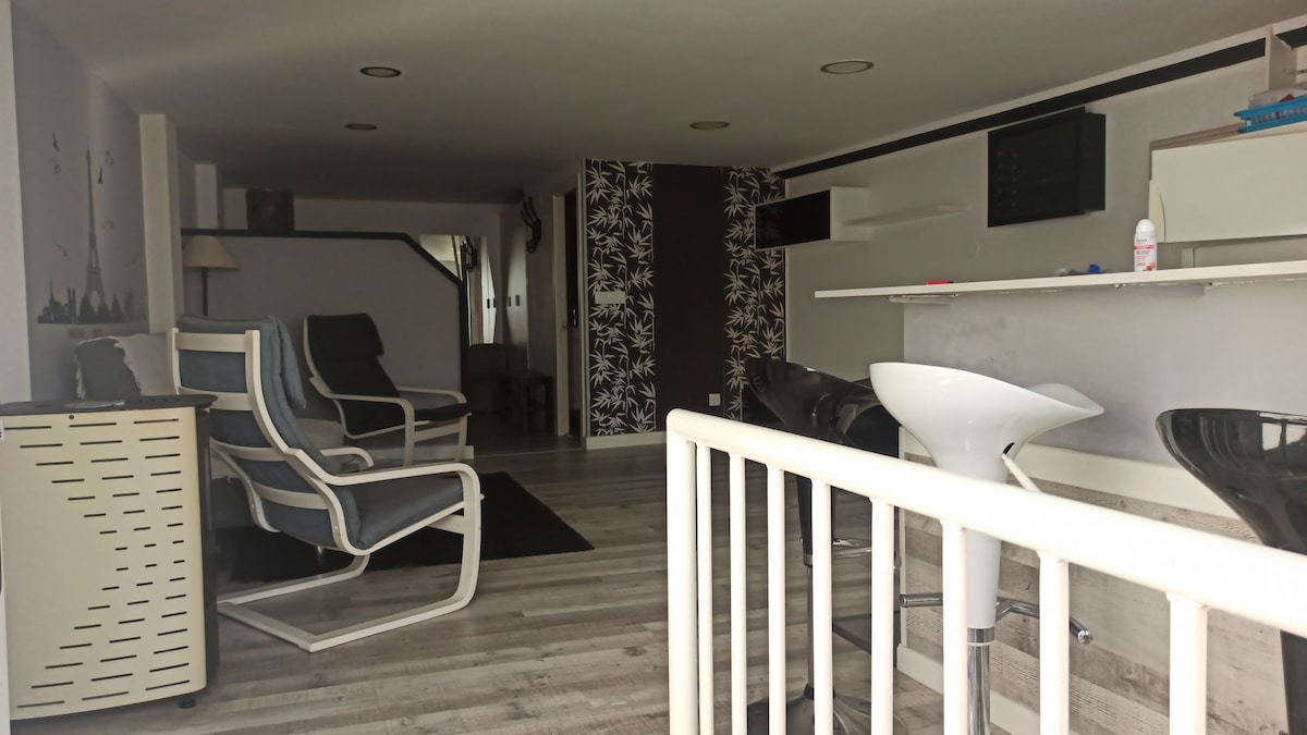 A modern living space features a light-colored floor and a variety of seating options, including two lounge chairs and a sleek bar area. Wall accents incorporate a floral design, while functional elements are subtly integrated into the open layout, providing a spacious ambiance.