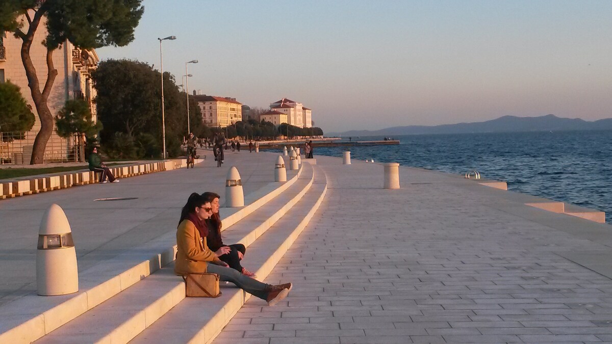 A spacious waterfront promenade extends along the coast, bordered by trees and buildings. Soft evening light casts a warm glow on the surface, while couples enjoy leisurely moments. Paved seating areas and decorative light fixtures are visible, enhancing the tranquil setting by the sea.