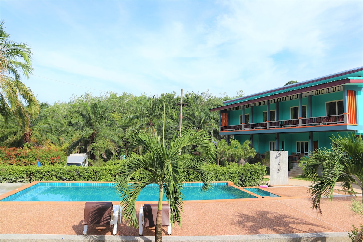 A serene pool area is surrounded by lush greenery, with a clear blue pool inviting relaxation. The two-story building features a vibrant exterior, complemented by the tropical landscape. Palm trees add to the natural ambiance of the space.