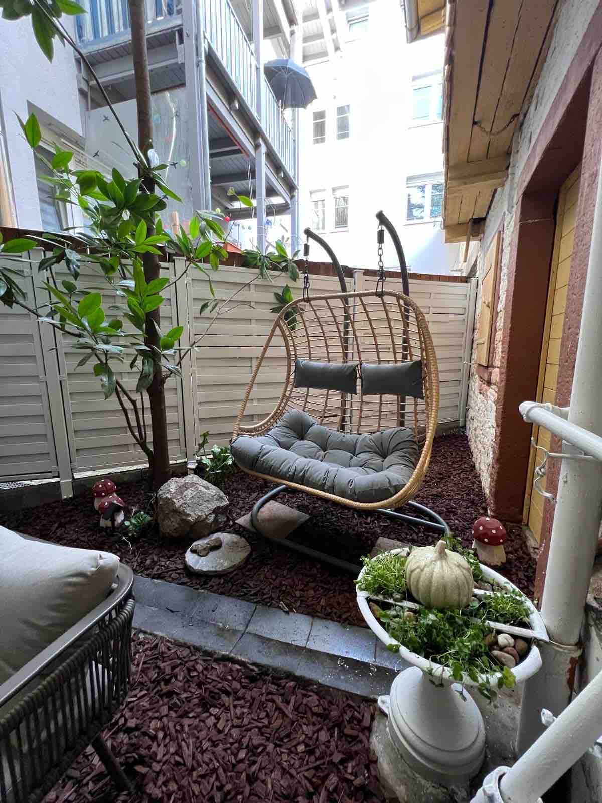 A cozy outdoor seating area features a hanging chair with soft cushions, surrounded by decorative stones and plants. The space includes a small garden area with low greenery and decorative elements like mushrooms and a charming pumpkin.