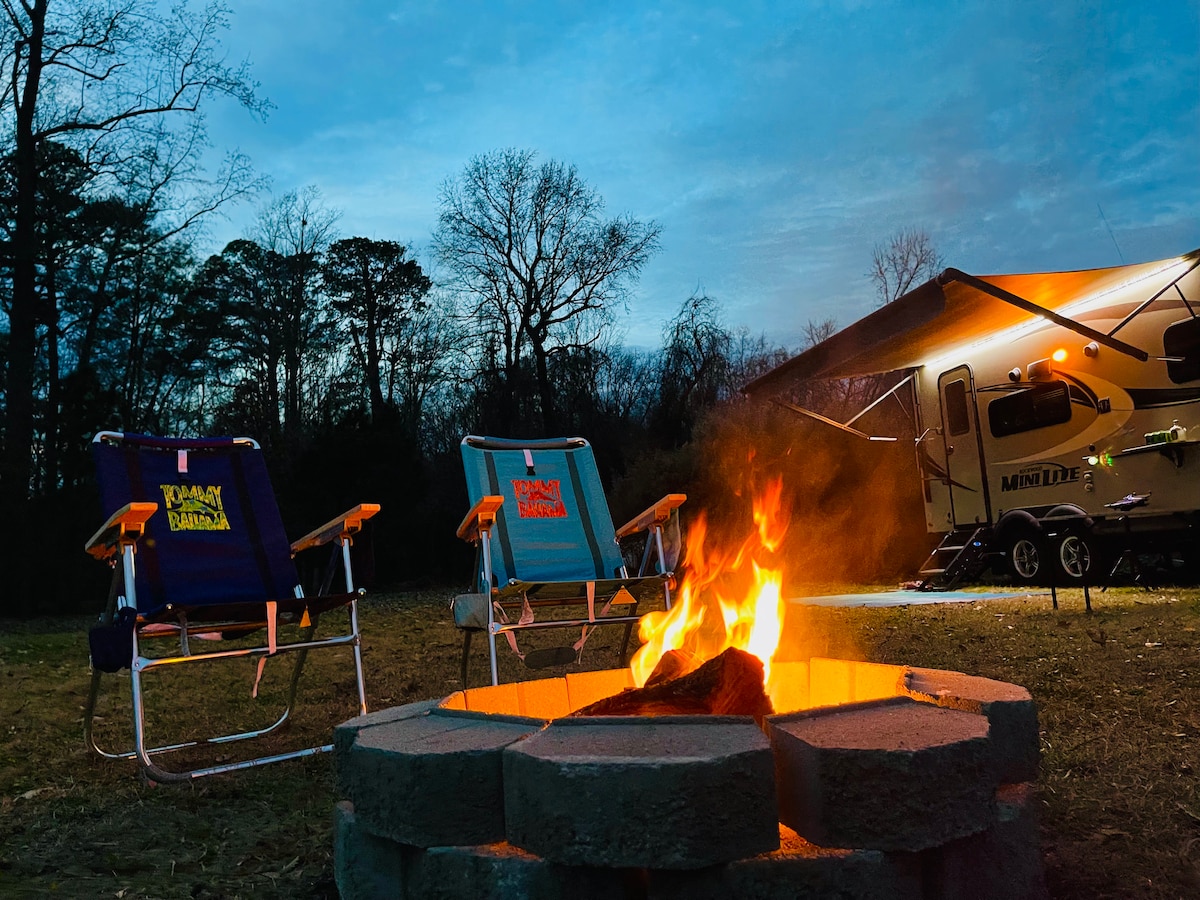 A cozy campfire is surrounded by a circular stone fire pit, with two folding chairs positioned nearby. A trailer is visible in the background, partially illuminated under the evening sky. Soft lighting from the trailer contrasts with the natural surroundings.