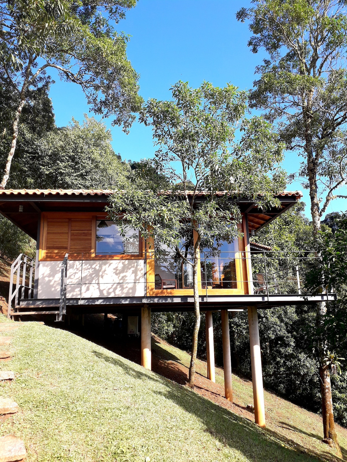 A modern chalet is positioned on a sloped landscape, elevated on stilts. Large windows allow natural light to fill the interior while offering views of the surrounding greenery. The structure features a wooden roof and a deck extending from the main entrance.