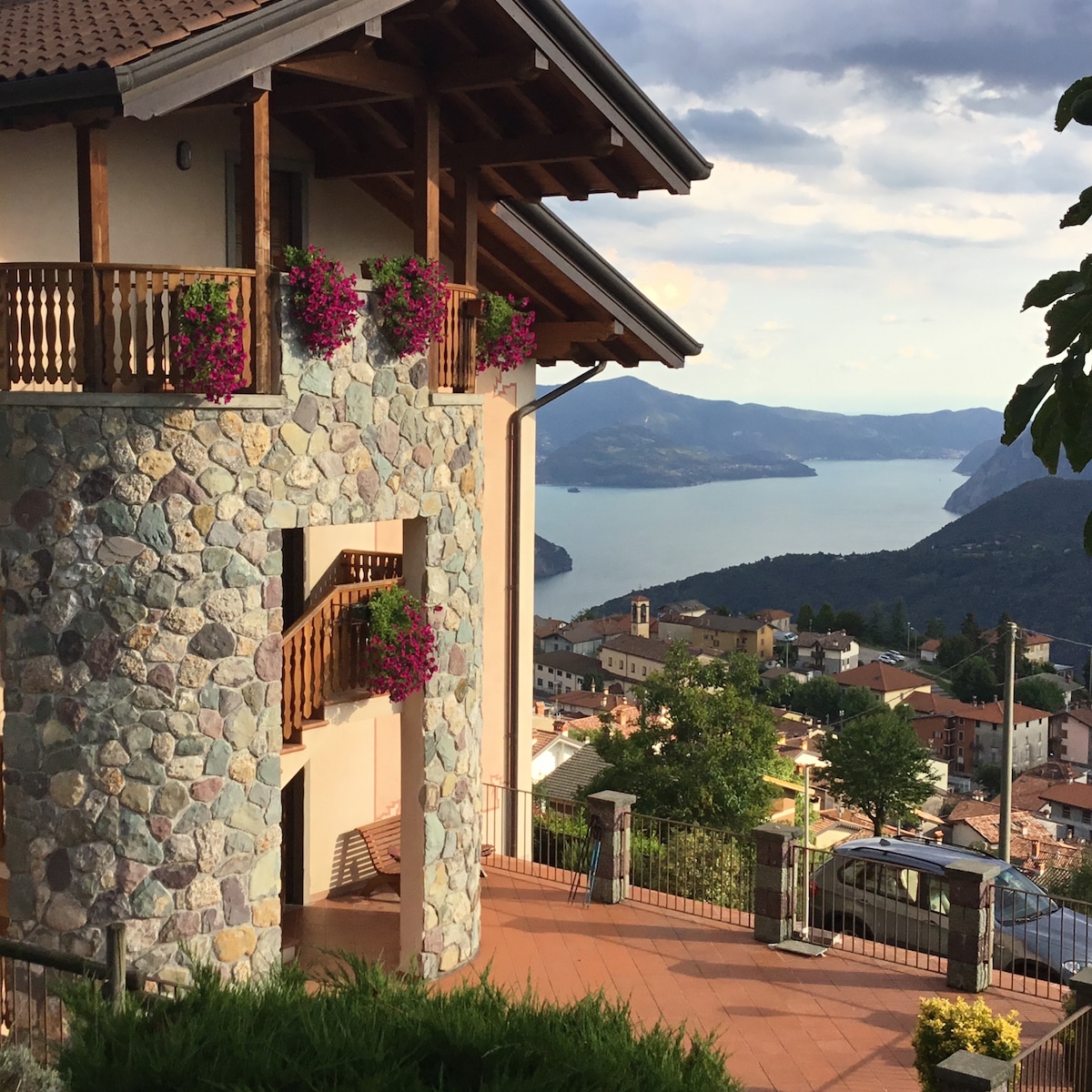 A stone exterior of a two-story villa is complemented by vibrant flower baskets hanging from the wooden balconies. The view extends to a scenic lake surrounded by mountains, with a hillside village visible in the foreground under a partly cloudy sky.