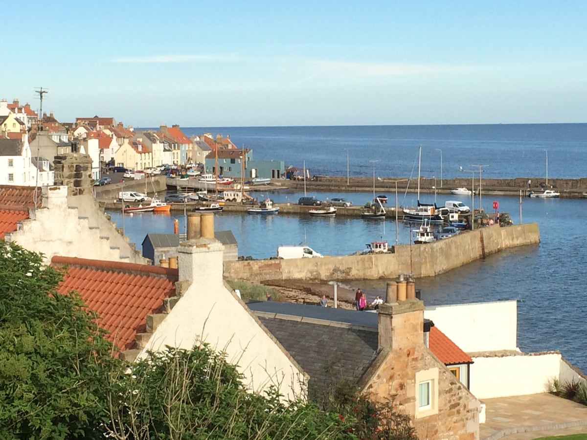A scenic view of the harbour features colorful houses lining the shore. Fishing boats are docked along the piers, with calm blue waters stretching into the distance. The backdrop includes a clear sky, creating a peaceful coastal atmosphere.