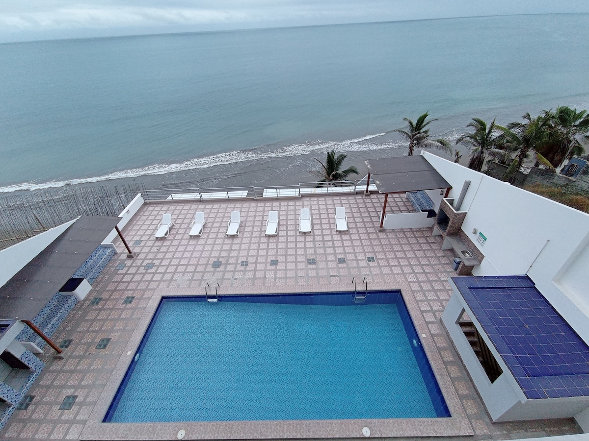 An aerial view of a pool area overlooking the ocean. A rectangular pool is surrounded by light-colored loungers, with palm trees visible along the shoreline. The area features shaded seating and a tiled deck, promoting a relaxing atmosphere with direct beach access.