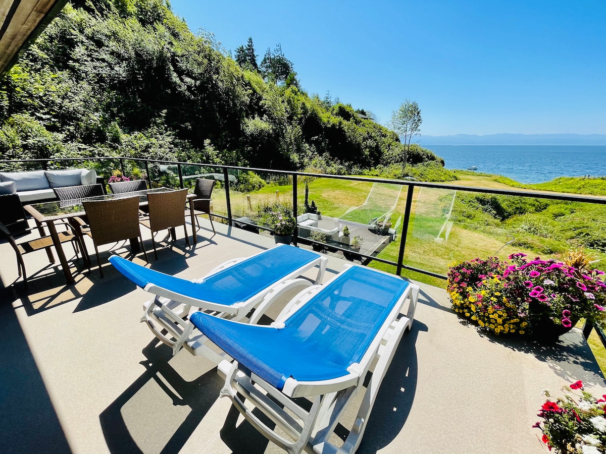 An outdoor patio features two lounge chairs with blue cushions, framed by a lush green lawn and colorful flower beds. A dining table with chairs and a barbecue area are visible, overlooking the ocean and a clear blue sky.