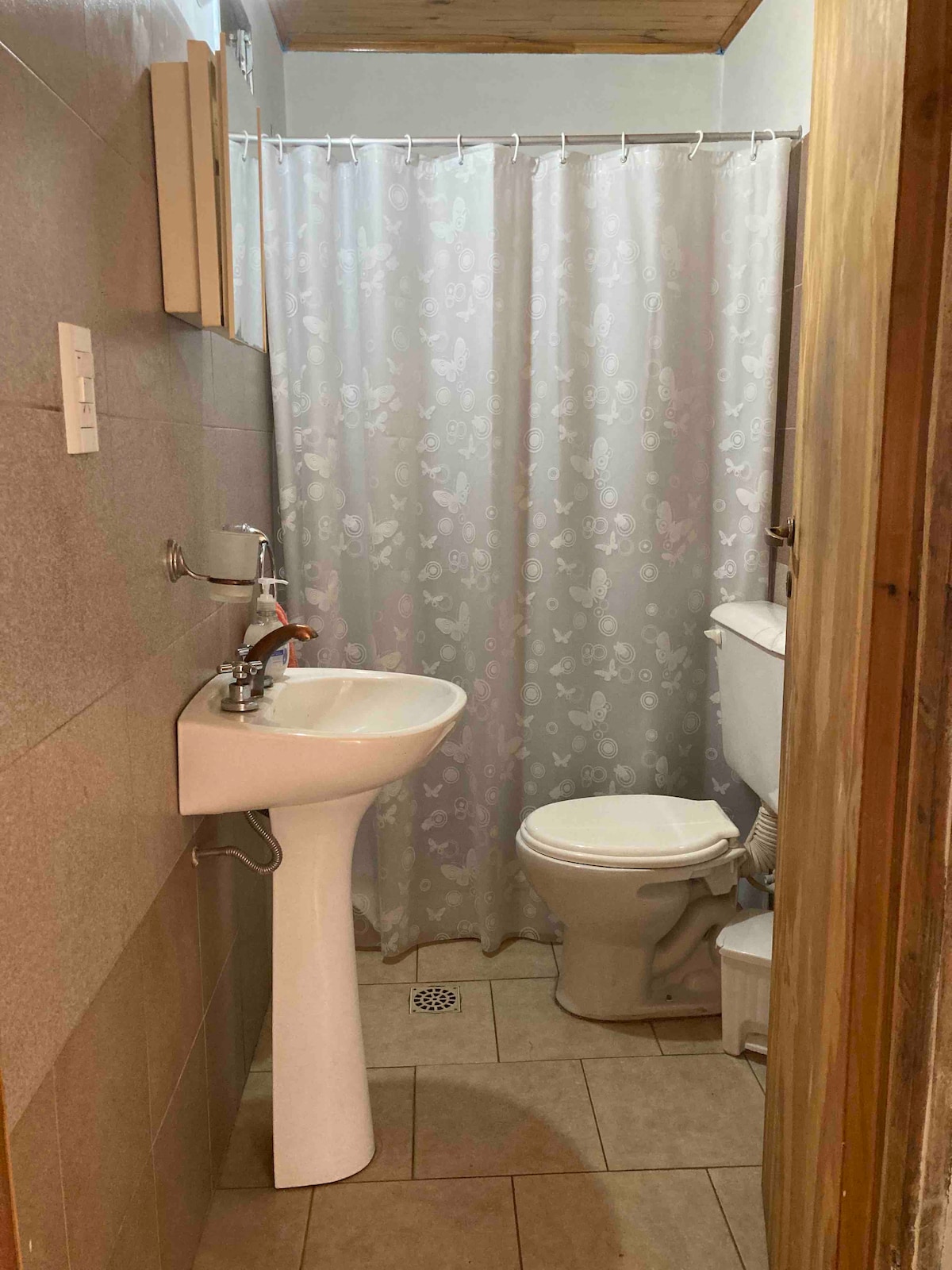 A compact bathroom features light-colored tile walls and a neutral floor. A white pedestal sink and a toilet are positioned next to each other, with a shower area enclosed by a floral-patterned curtain.