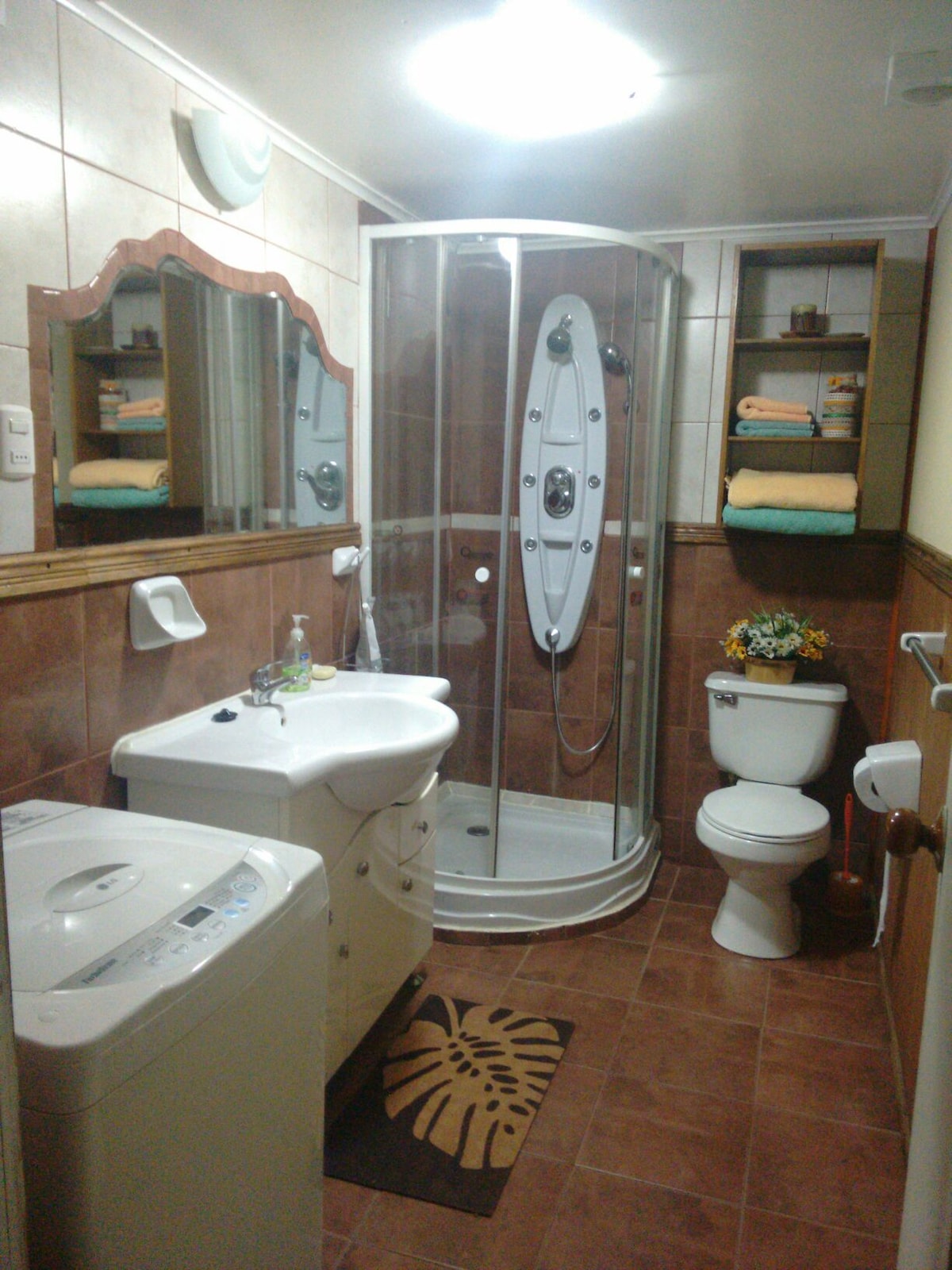 A clean bathroom features a circular shower stall with glass doors, a white sink, and a toilet. A washing machine is positioned beside the sink, while shelves hold neatly arranged towels. Warm lighting enhances the tiled walls and floor, contributing to a functional space.