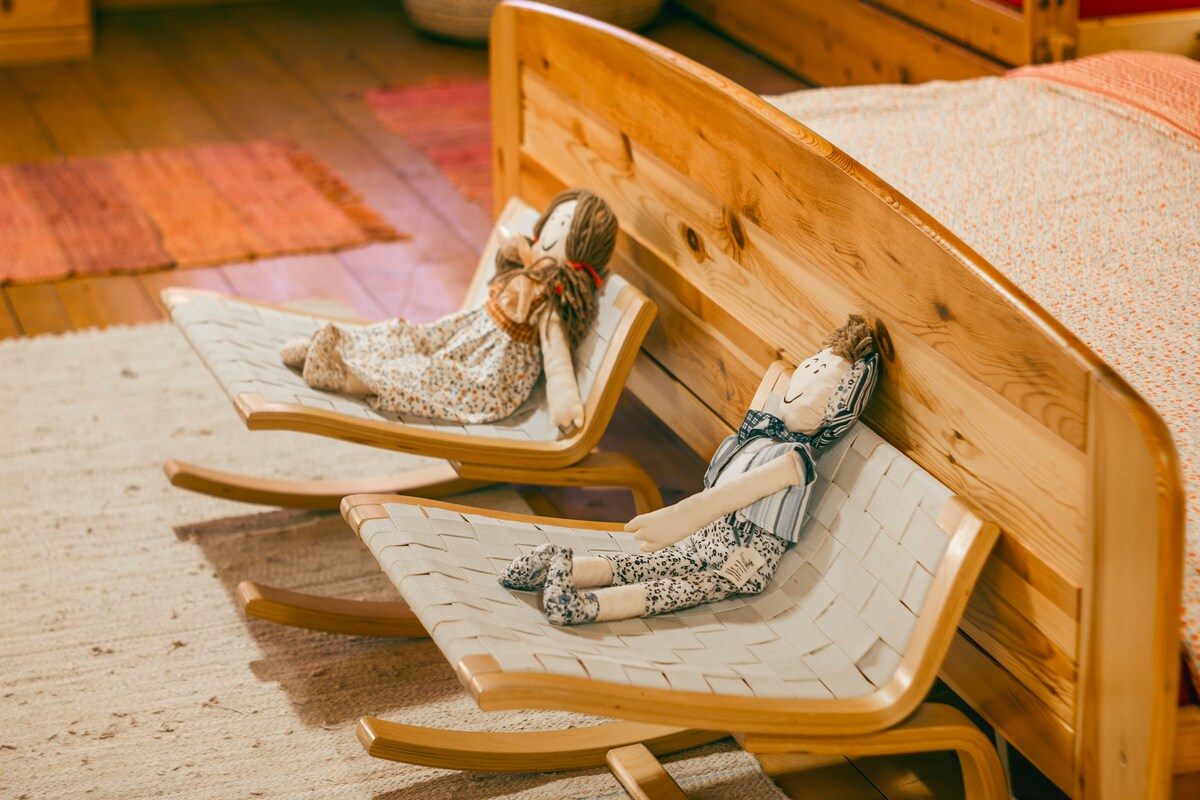 Two handmade dolls are positioned comfortably on small rocking chairs beside a wooden bed. The dolls, dressed in patterned clothing, contribute a charming touch to the room's warm wooden tones and textures, enhancing the inviting atmosphere.