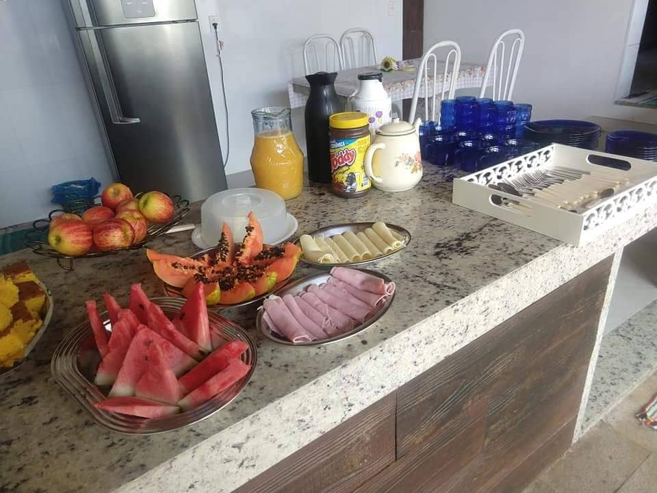 An assortment of fresh fruits and snacks is displayed on a countertop. Plates contain slices of watermelon, papaya, and various rolls. Glass containers hold beverages, and the background features a neat arrangement of dishes. Colorful bowls and utensils are visible, adding to the inviting setup.