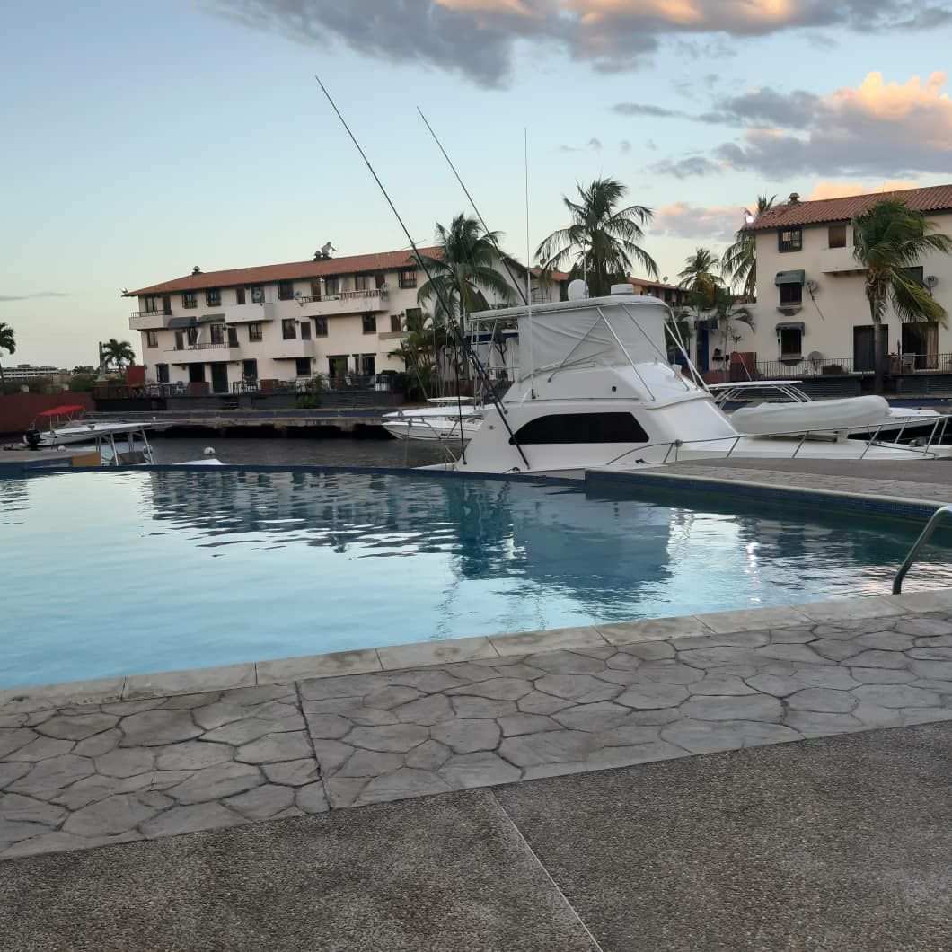 A pool area is visible, featuring calm water reflecting the sky. Several boats are docked nearby, with palms swaying in the background. Two-story buildings line the waterfront, providing an inviting scene for relaxation.