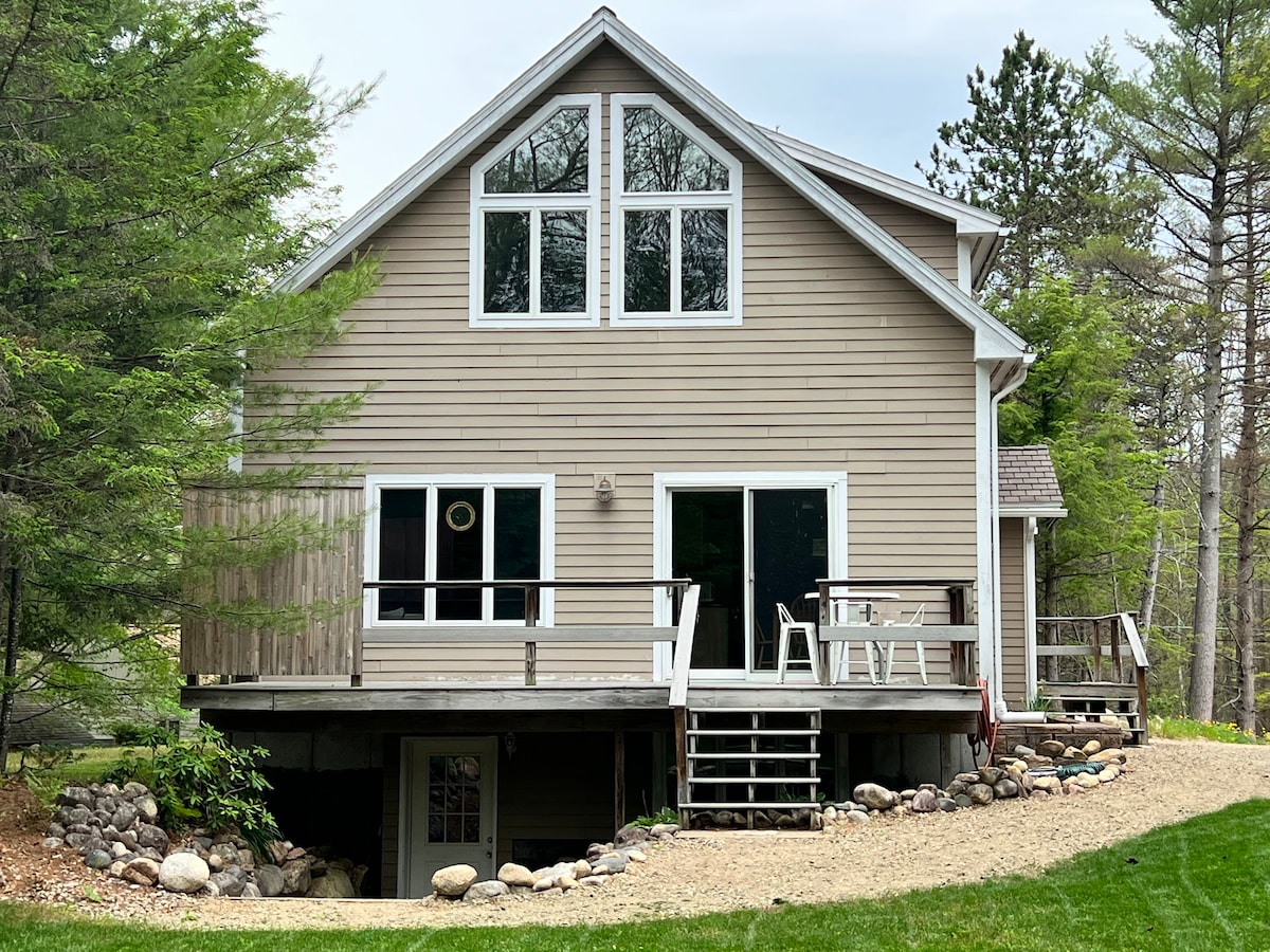 The exterior of a two-story home is presented, showcasing a large deck with multiple seating options. Large windows allow natural light to fill the interior. Surrounding greenery and a gravel path lead to the entrance, while a stone border adds texture to the landscape.