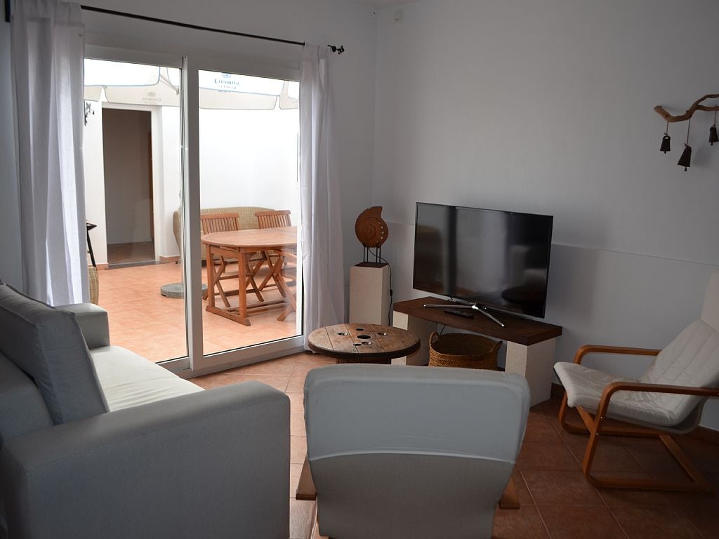 A light-filled living area displays a comfortable seating arrangement with two sofas and a lounge chair. A large television is positioned on a stand, facing the seating. Floor-to-ceiling glass doors open to a patio, allowing natural light to flow into the space.