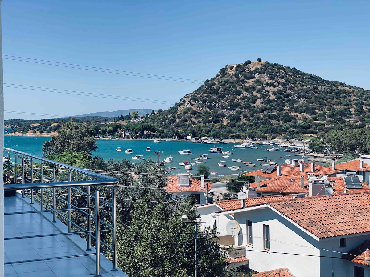 A spacious balcony provides a view of a serene harbor with small boats gently bobbing in the turquoise water. The landscape highlights lush green hills in the background, complemented by the charming rooftops of nearby buildings.