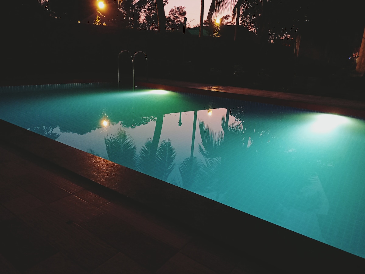 The swimming pool is illuminated at night, showcasing clear water with gentle turquoise hues. Reflections of palm trees and soft light create a serene ambiance. The pool area is surrounded by greenery, emphasizing a peaceful retreat.