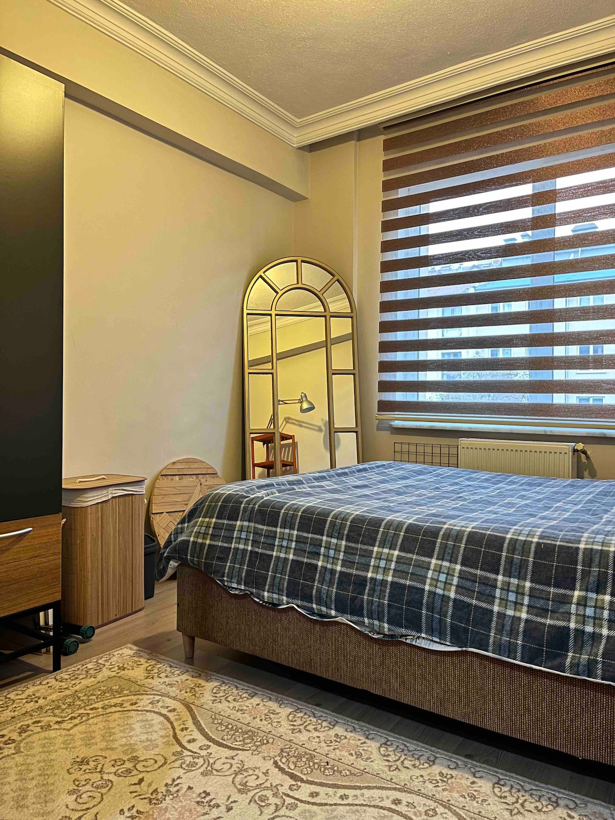 A cozy bedroom features a spacious bed covered with a plaid blanket. A tall mirror leans against the wall, reflecting natural light. A small chair and a wooden side table are positioned in the corner, with subtle decorative elements enhancing the environment.