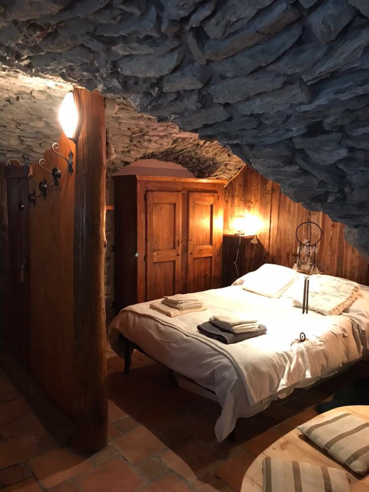 A cozy sleeping area is designed with rustic stone walls and wooden accents. A spacious bed with fresh linens is situated beneath a soft glow from a nearby lamp. An armoire provides ample storage, complimenting the overall warm aesthetic of the space.