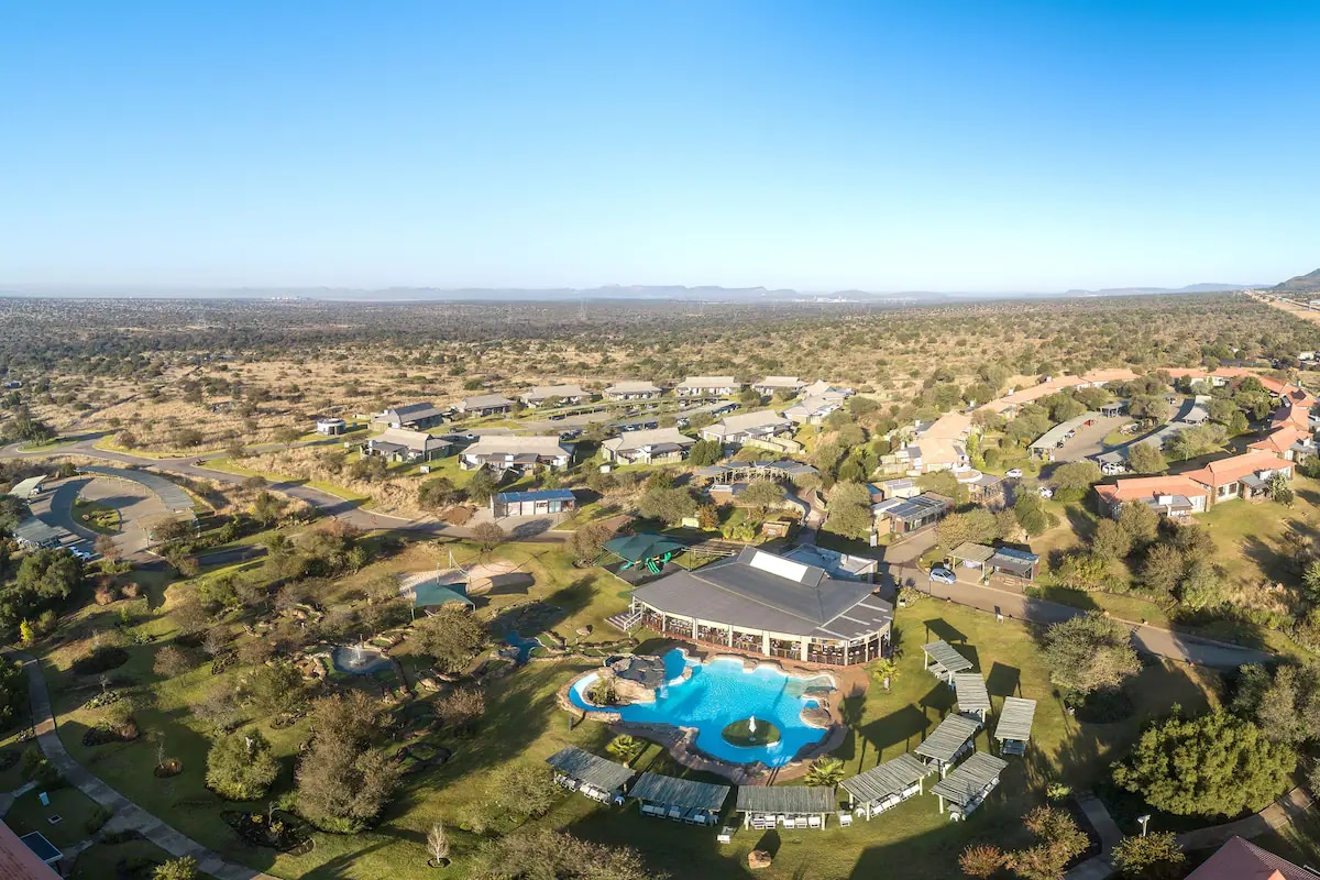 An aerial view captures the resort's layout, featuring a central building surrounding outdoor amenities. A sparkling pool is visible, accompanied by shaded lounging areas. Lush greenery and pathways enhance the natural surroundings, providing a glimpse of the expansive landscape extending into the distance.