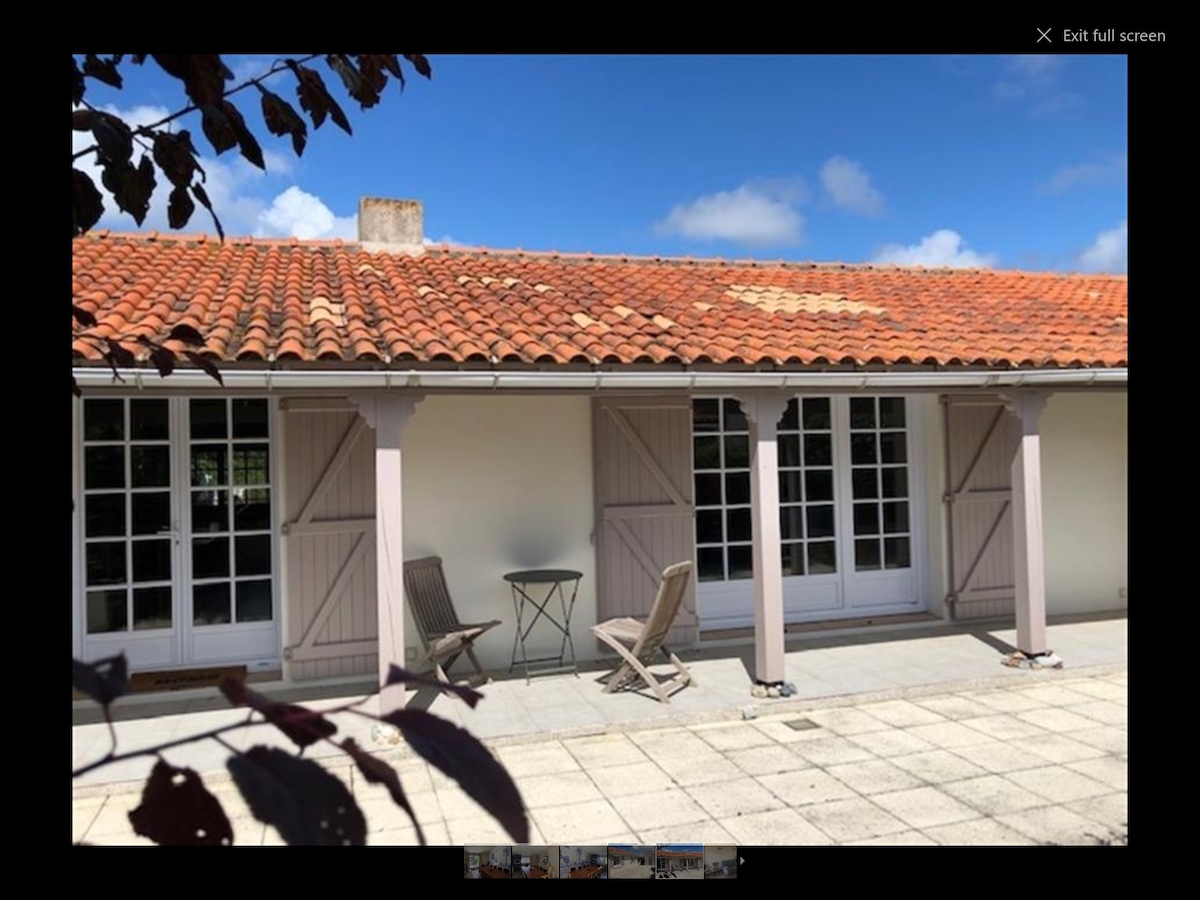 The exterior of the villa features a terracotta roof and cream-colored walls. Two sets of French doors, framed by wooden shutters, open to the outdoors. A small, round table is flanked by two wooden chairs on the tiled patio, under a clear blue sky.