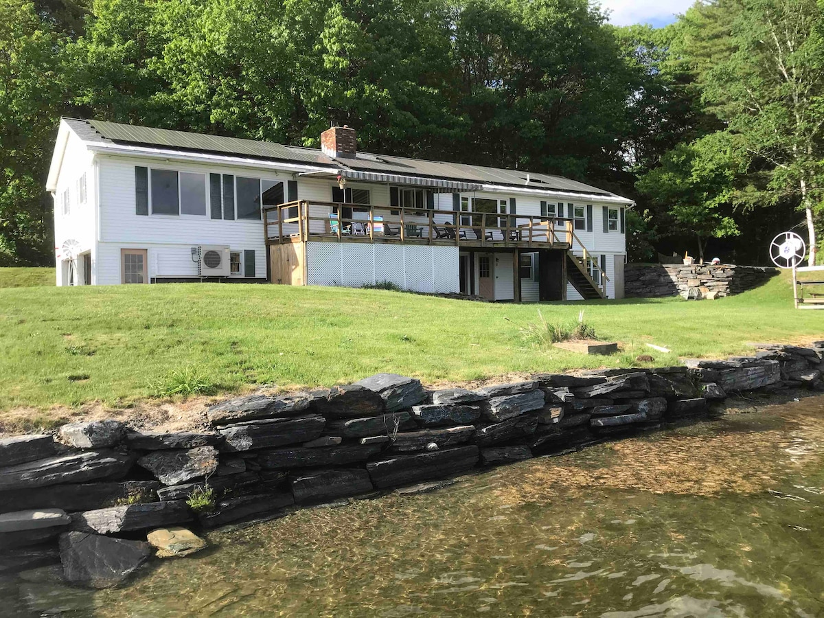 The lakefront home features a wide covered deck and large windows, providing expansive views of the surrounding greenery. The structure has a white exterior and a stone foundation, set against a well-maintained lawn leading to the water's edge.