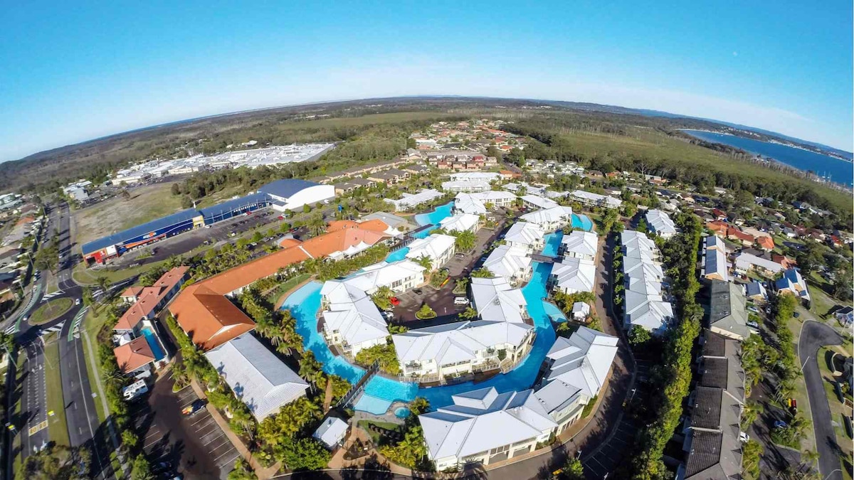 An aerial view reveals the layout of the Oaks Pacific Blue Resort, showcasing a series of white-roofed villas with a prominent lagoon pool winding through the property. Surrounding greenery enhances the scene, while the nearby coastal area and roads can be seen in the background.