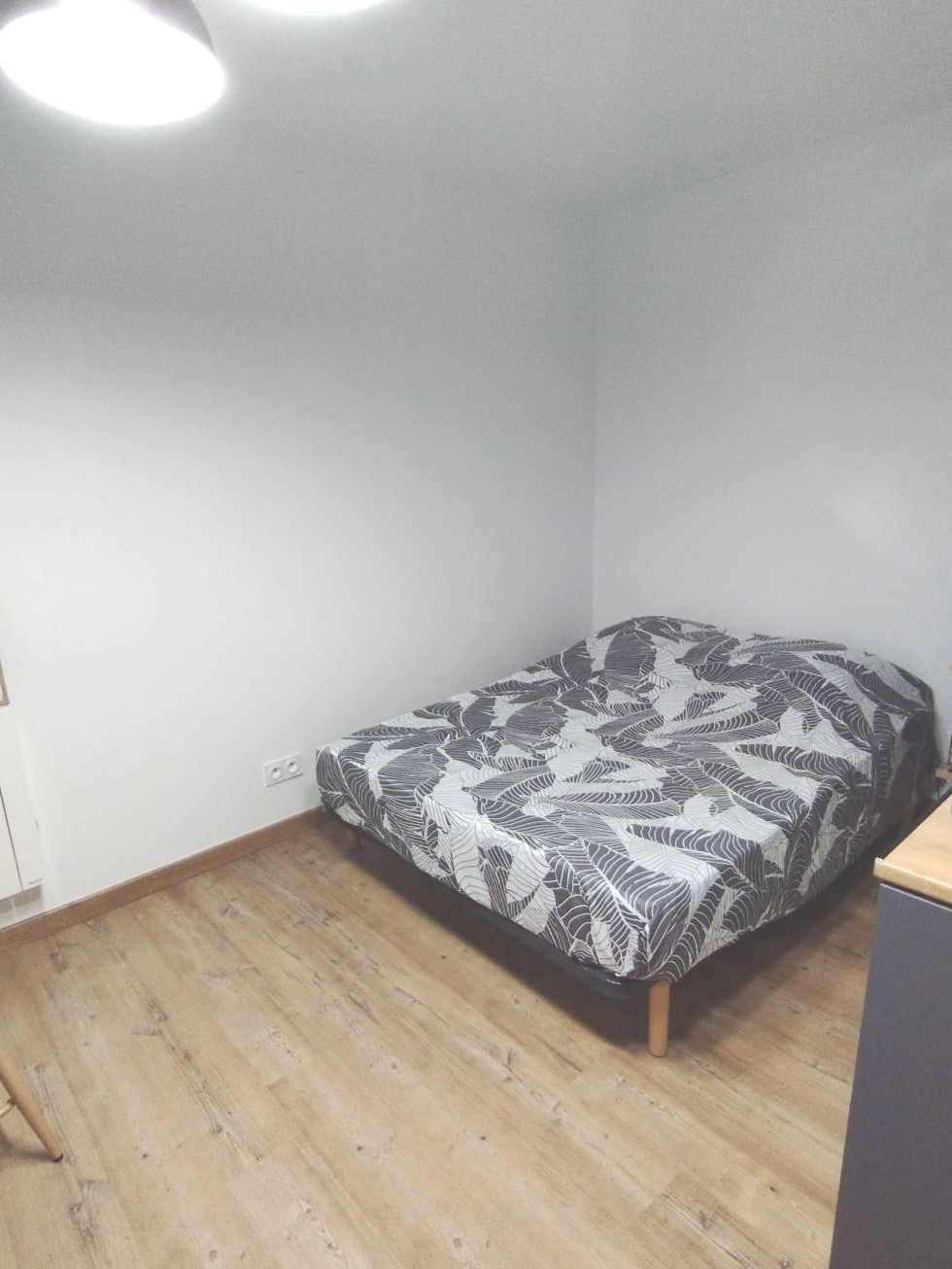 A simple sleeping area features a bed with a patterned duvet cover against a light wall. Wooden flooring is visible, along with a small bedside table positioned nearby. Soft overhead lighting creates a calm ambiance in the space.