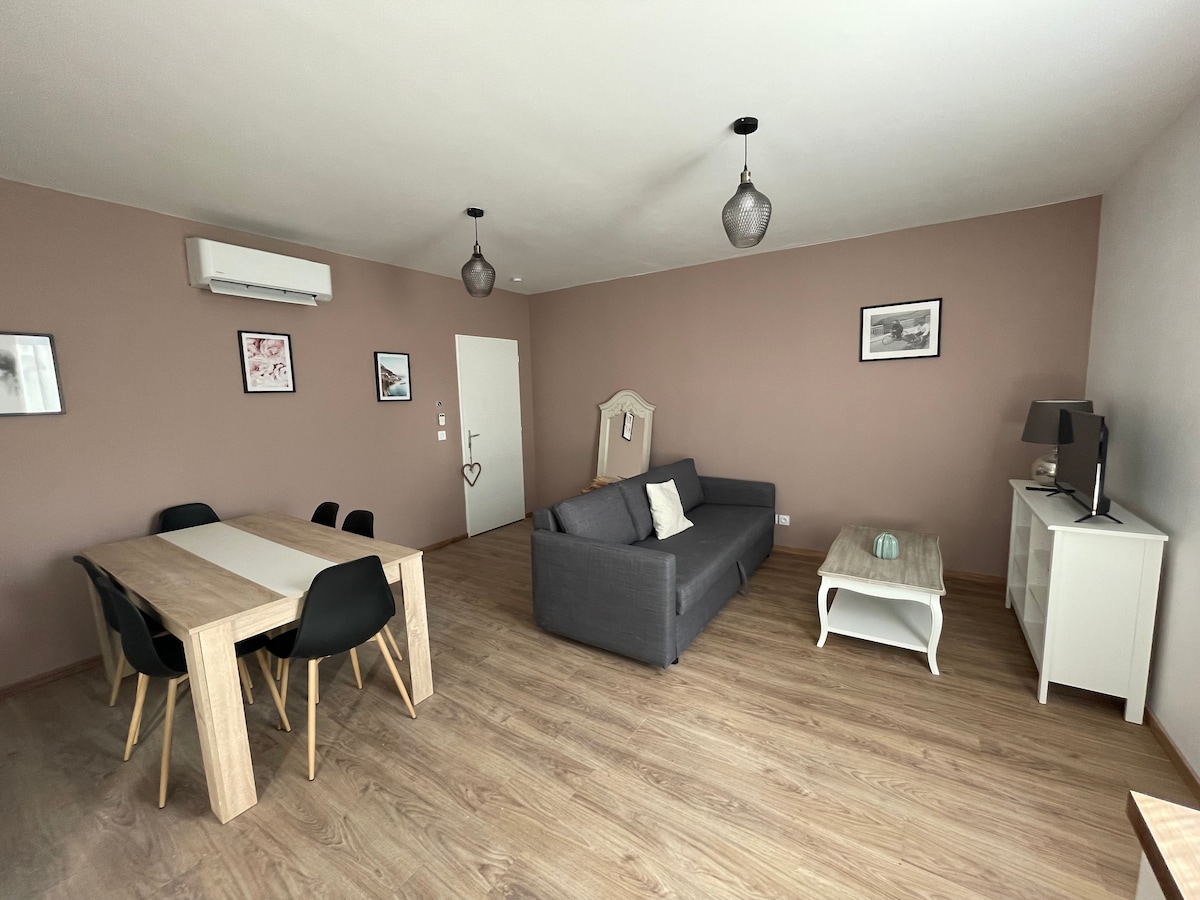 A spacious living area is presented, featuring a gray convertible sofa and a light wooden dining table surrounded by black chairs. Wall-mounted lamps provide soft lighting, and framed artworks adorn the neutral-colored walls, creating an inviting atmosphere.