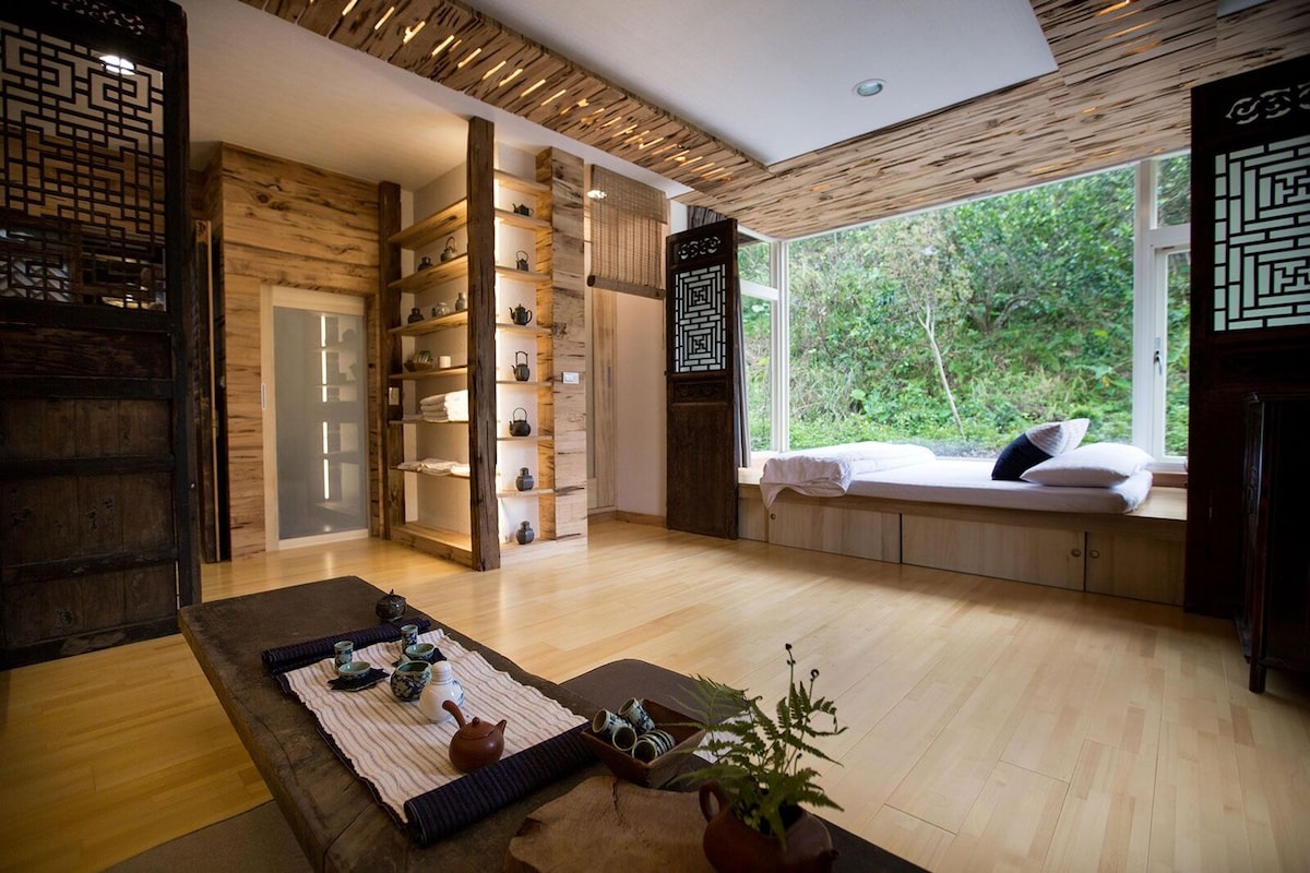 A serene interior space features wooden accents and natural light streaming through large windows. A low table is set with tea utensils, while a cozy bed is positioned against the window with soft bedding. Shelving displays decorative items, enhancing the calm ambiance.