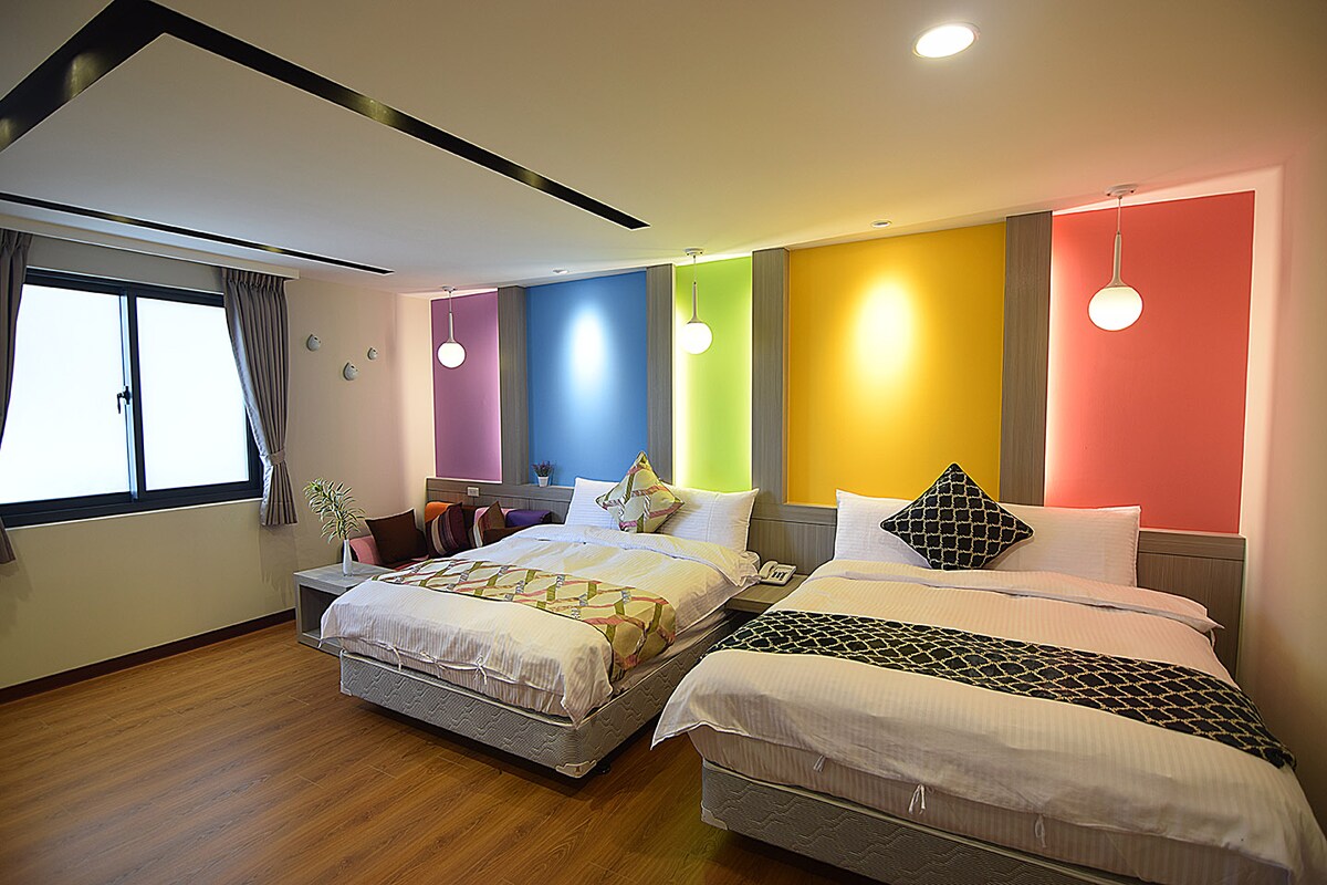 A bright sleeping area features two spacious beds, one adorned with a patterned blanket. Colorful accent walls in shades of blue, green, yellow, and pink enhance the modern design. Large windows with curtains allow natural light to fill the room, complemented by minimalistic pendant lighting.