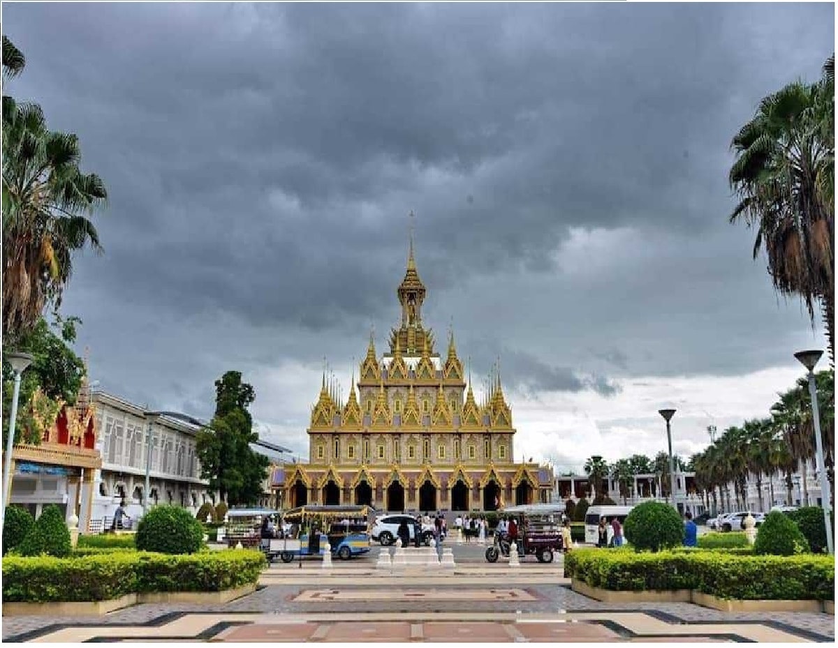 An ornate building featuring intricate golden architecture stands under a cloudy sky. Lush green landscaping surrounds the structure, with palm trees framing the scene. Pathways lead towards the entrance, where small vehicles are parked, inviting visitors to explore.