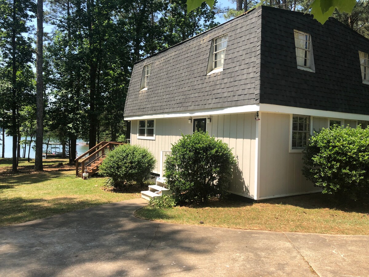 A charming two-story house is framed by lush greenery and tall trees. A sloped driveway leads to the front entrance, where wooden steps ascend to the door. The exterior features a mix of light siding and a dark, shingled roof, creating a warm contrast with the natural surroundings.